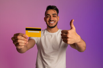 Happy young Arab man holding credit card and showing thumb up gesture in neon light, selective focus