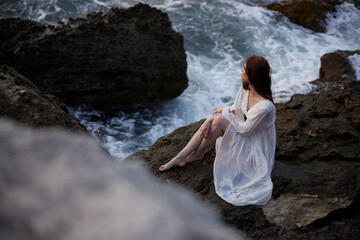 A woman in a white dress barefoot sits on a stone on a cliff top view
