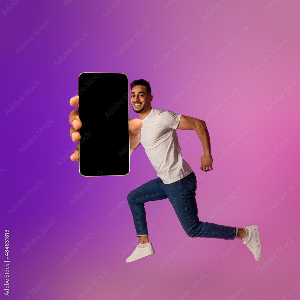 Poster full length portrait of happy young arab guy jumping with smartphone, showing empty screen in neon l