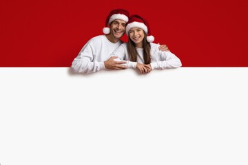 New Year Ad. Happy couple wearing santa hats leaning at blank board