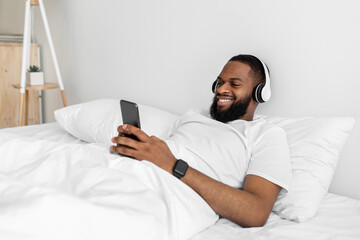 Cheerful young african american man with beard in headphones, typing on smartphone, enjoy listening music