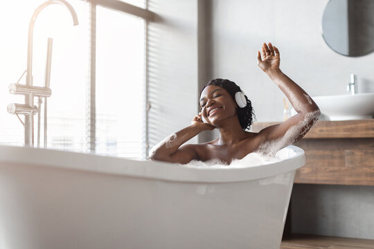 Positive african american woman taking foamy bath and listening music