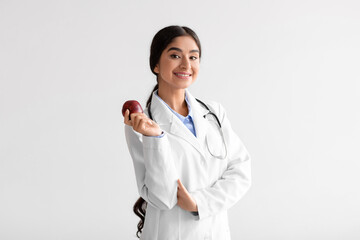 Happy pretty young indian woman doctor dentist in white coat with stethoscope hold fresh red apple