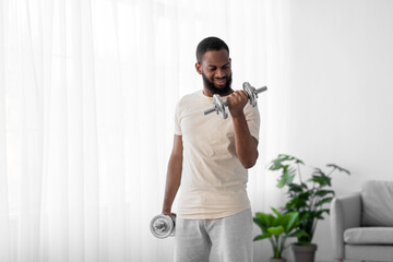 Cheerful millennial bearded african american guy in white sport wear lifts dumbbells in room interior near window