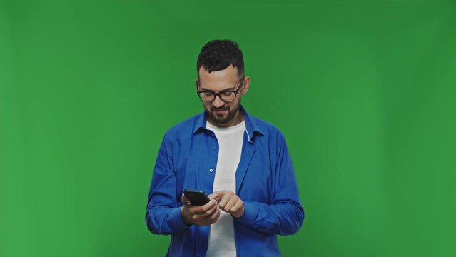 Smiling handsome bearded mand in casual shirt isolated on green background studio. Using mobile phone typing sms message browsing