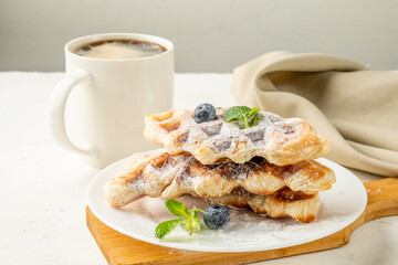 Croffles in a light plate sprinkled with powdered sugar and blueberries, on a cloth background and a mug with black coffee. A mixture of curassan and waffles.