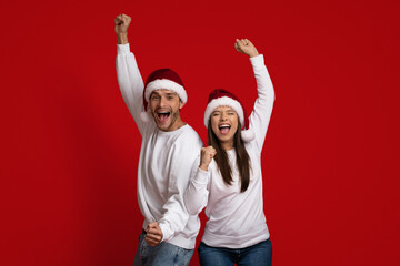 Fototapeta premium Christmas Celebration. Overjoyed Couple In Santa Hats Celebrating Success With Raised Fists