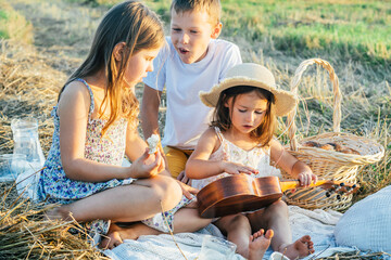 Portrait of joyful boy and two girls sitting on blanket in field, having picnic, eating bread....