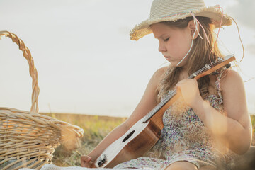 Portrait of girl sitting on blanket in dry hay field, having picnic, learning playing ukulele....