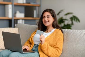 Happy arab woman with laptop and credit card shopping online, using web banking service, sitting on sofa at home