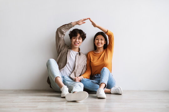 Asian couple making house roof, joining hands above heads as symbol of new home, sitting on floor against white wall