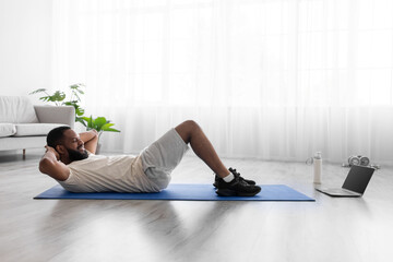 Smiling young black man with beard in white sportswear do abs exercises and watch video on laptop on floor