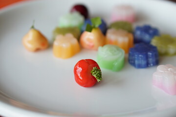 Close-up of strawberry fruit shape Thai dessert is Look Choup with Kanom Chan under sunlight and white plate on wood table.