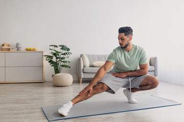 Fit millennial Arab guy stretching his leg, working out at home during covid lockdown, copy space