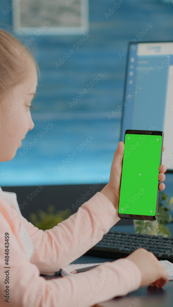 Poster child vertically holding smartphone with green screen while sitting at desk and doing homework. litt