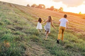 Portrait of two girls in dress and boy back to camera, playing game of catch and running on grass hay field paths of dry grass in the sunset. Forest on background. Cloudy sunny sky. Haying time