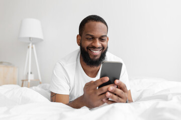 Obraz na płótnie Canvas Happy millennial african american bearded man lies on white bed in bedroom interior and typing on smartphone