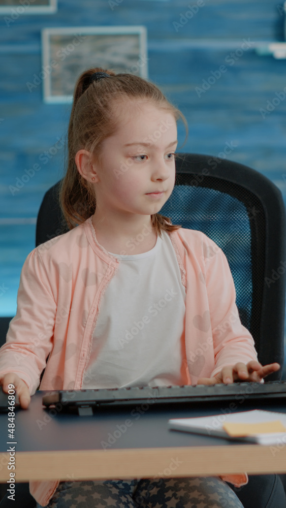 Poster little girl putting backpack on couch and using computer with keyboard at desk. young child looking 