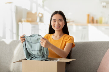 Mail delivery, shipment concept. Excited asian woman unpacking parcel, holding new shirt, satisfied with purchase