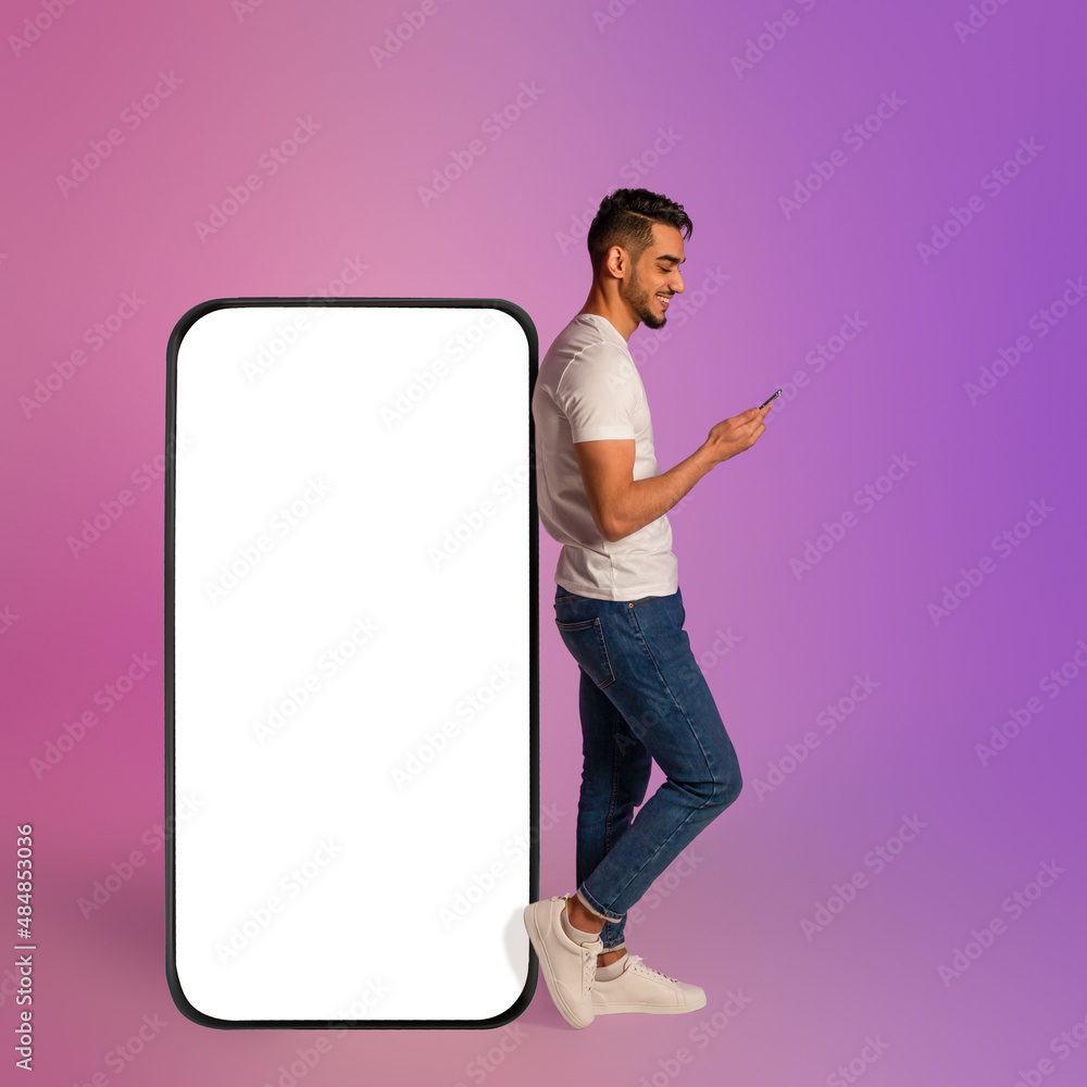 Poster millennial arab man using mobile device while leaning on giant smartphone with blank white screen in