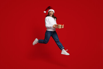 Fototapeta na wymiar Positive Millennial Woman Wearing Santa Hat Jumping In Air With Gift Box