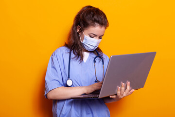 Woman nurse with face mask looking at laptop and wearing uniform and stethoscope. Medical assistant using modern computer during pandemic, person standing over isolated background.