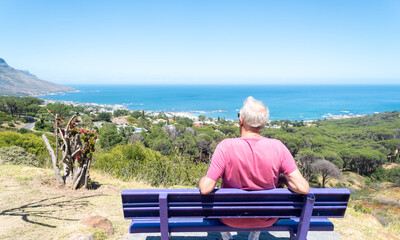 senior citizen, elderly man sits and rests on a bench looking at a scenic coastal view of Camps Bay beach, Cape Town, South Africa