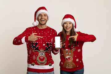 Mobile Ad. Cheerful Young Couple Wearing Santa Hats Pointing At Blank Smartphone
