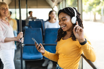 Cheerful black lady using smartphone wearing headphones in city bus