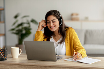 Smiling young arab woman wearing wireless headset and using laptop, watching video or having video...