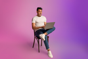 Shocked young Arab guy looking at laptop screen, sitting on chair, excited about online win or success in neon light - obrazy, fototapety, plakaty