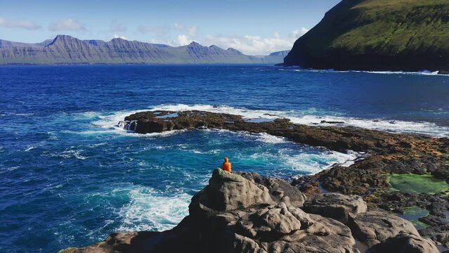 Aerial view of seascape. Man siting and looking beautiful landscape, waves crashing on rocky coast. Rough sea and mountain, cliff in background during summer sunny day.