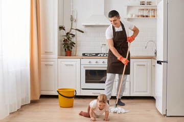 Indoor shot of brunette man wearing brown apron washing floor in kitchen, cleaning flat with hisi nfant baby daughter, helping his wife doing work about the house.