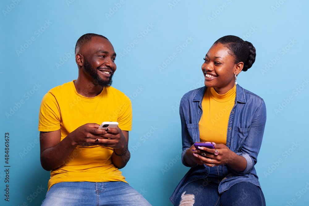 Poster boyfriend and girlfriend holding mobile phones and smiling at each other, using smartphones with tou