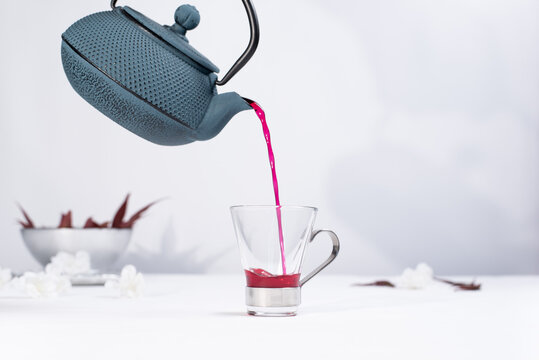 Verter te rojo de bayas en una taza transparente con tetera de hierro azul sobre una mesa blanca y un fondo gris claro	