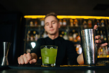 Close-up low-angle view of confident bartender male giving ready cocktail to customer standing behind bar counter in modern nightclub, on blurred background of shelves with different alcoholic drinks.