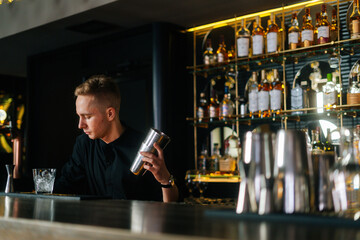 Side view of professional bartender preparing refreshing alcoholic cocktail standing behind bar counter in modern dark nightclub, on blurred background of shelves with different alcoholic drinks.