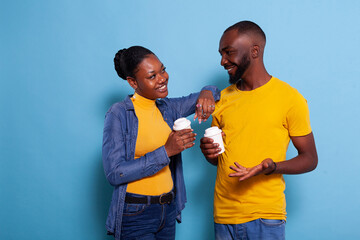 Obraz premium Romantic couple in love looking at each other in studio, holding cup of coffee in hand. Girlfriend and boyfriend flirting and smiling, standing over blue background and enjoying drink.