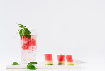 Un vaso de jugo de sandía con hojas verdes trozos de sandia y hielo sobre fondo claro	