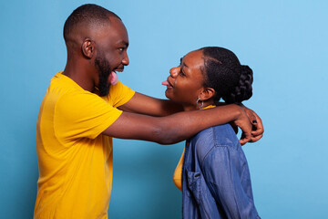 Playful lovers hugging and sticking tongue out at each other. Girlfriend and boyfriend fooling around with goofy gesture, expressing happiness and corny romance. Positive couple having fun.