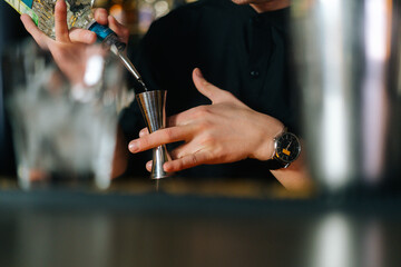 Fototapeta na wymiar Close-up cropped shot of unrecognizable bartender male pouring alcoholic beverage into metal jigger, preparation cocktail with liquor standing behind bar counter in modern nightclub with dark interior