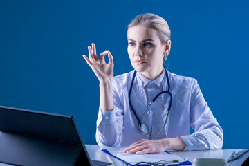 Hospital. A female doctor sits at a desk on a blue background, shows ok. Chief Physician. Therapist