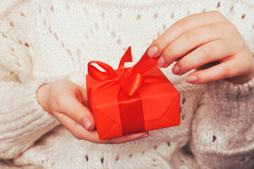  Close up shot of female hands holding a gift wrapped with red ribbon. Woman in white or beige...