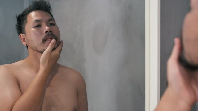 Adult man shaving with foam and manual razer.Mirror reflection young handsome smiling man touching beard, grooming in bathroom. Head shot close up happy asian man guy doing morning beauty routine.