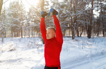 Outdoor winter sports. Athletic senior man working out with dumbbells, training arm muscles at snowy forest