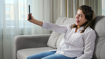 Cheerful disabled woman with cerebral palsy takes selfie via mobile phone smiling and making gestures sitting on comfortable sofa at home. - obrazy, fototapety, plakaty