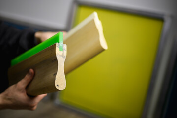 Partial shot of craftsman holding screen printing squeegees near blurred frame in workshop 
