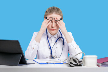 A female doctor sits in glasses at her desk near a laptop on a blue background, feels a headache...