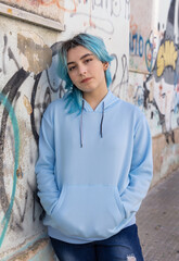 Blue haired Teenage girl in light blue oversize hoodie pointing against graffiti wall