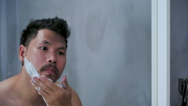 Adult man shaving with foam and manual razer.Mirror reflection young handsome smiling man touching beard, grooming in bathroom. Head shot close up happy asian man guy doing morning beauty routine.